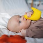 Cute newborn with toy duck in mouth lying on bed